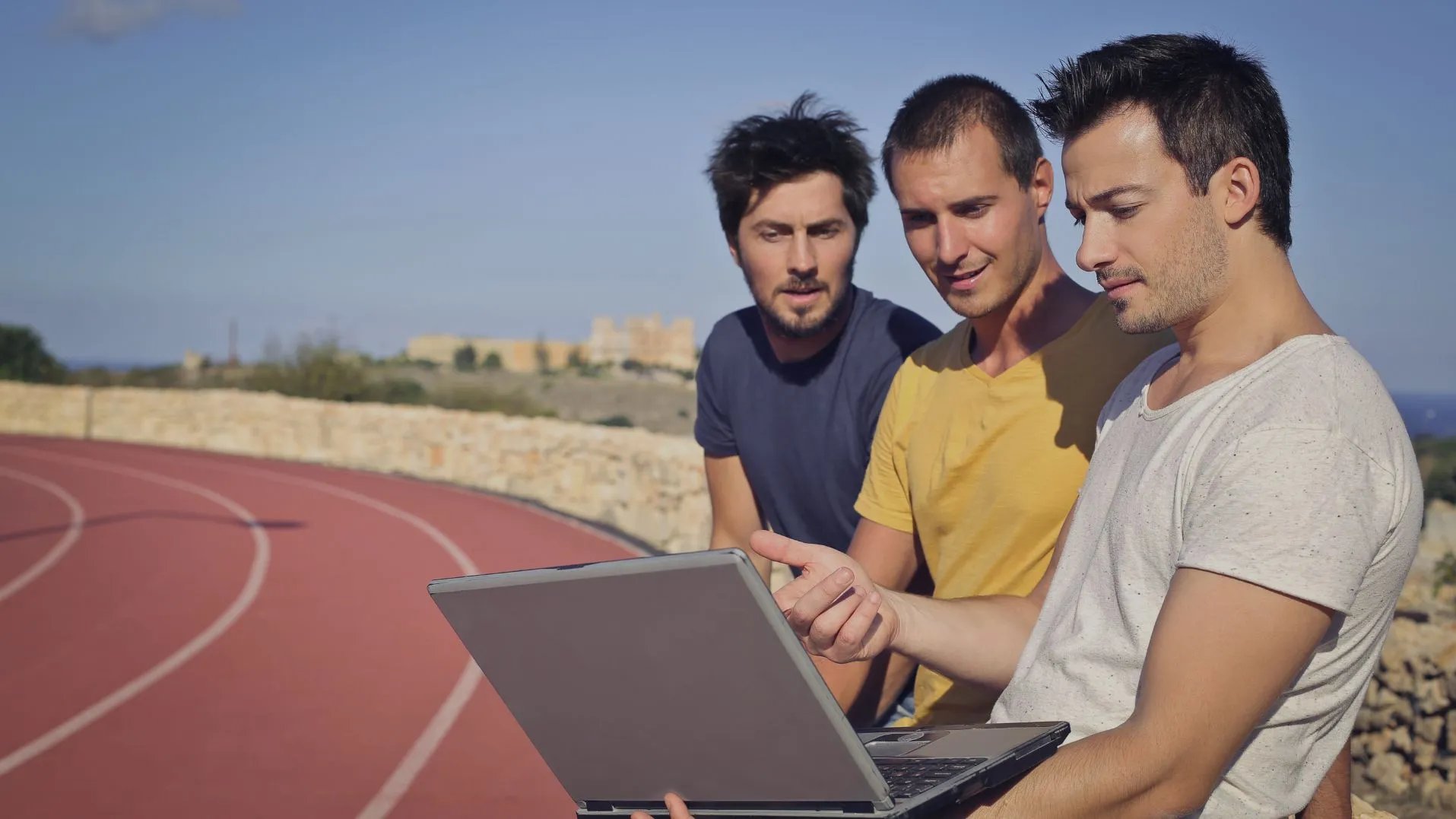 Drei Männer sitzen am Rand einer Laufbahn vor einem Computer